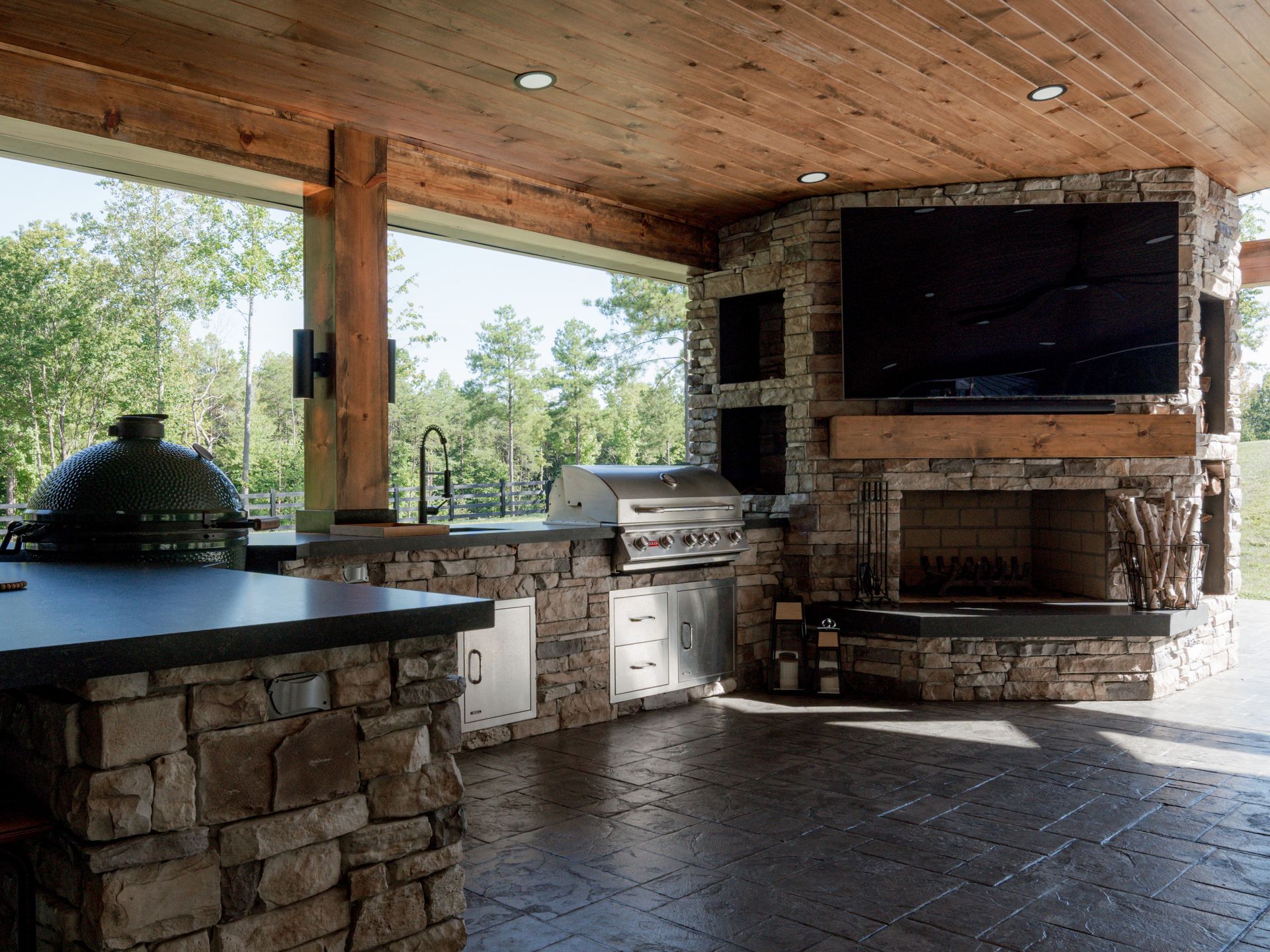 Stone outdoor kitchen and patio with grill, fireplace, and large TV under a wooden roof