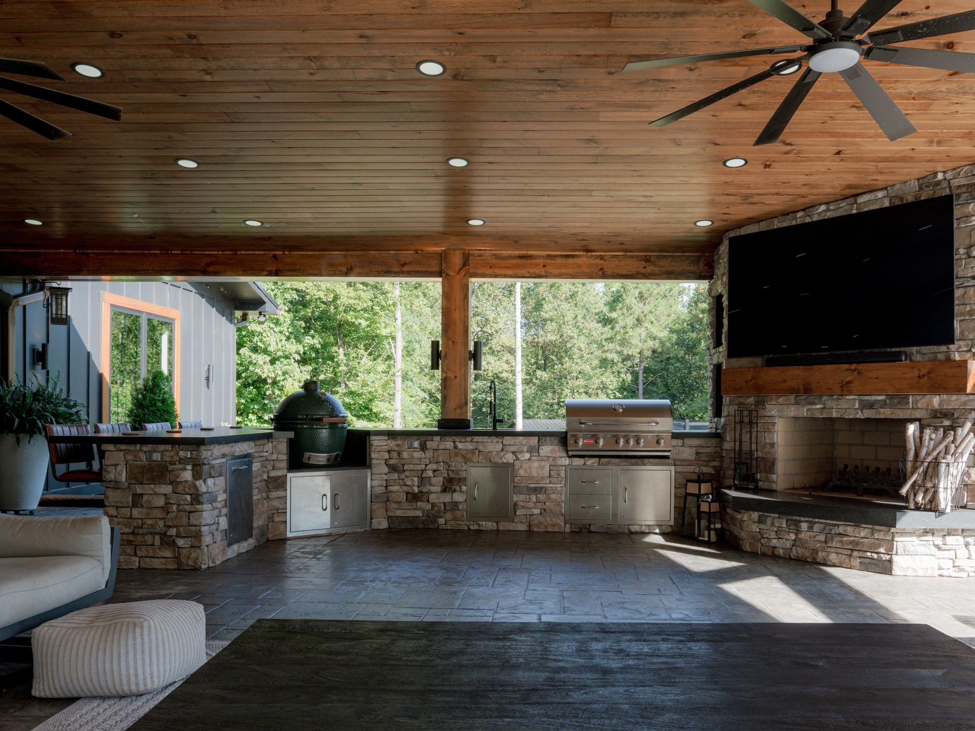 Covered outdoor patio with stone bar, wooden ceiling, ceiling fan, and mounted TV.