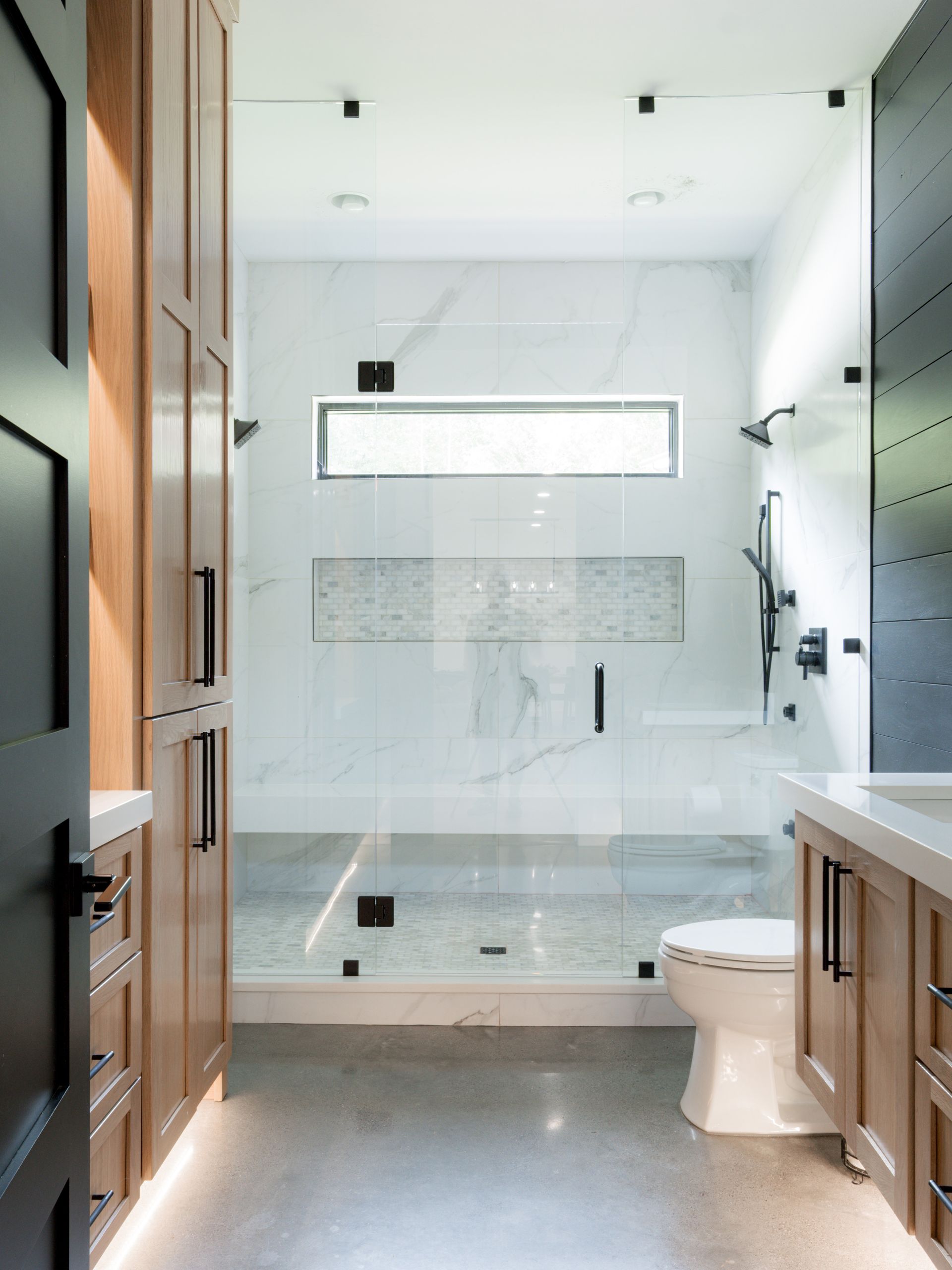 Bright modern bathroom with glass shower, white tile, window, and wooden door.