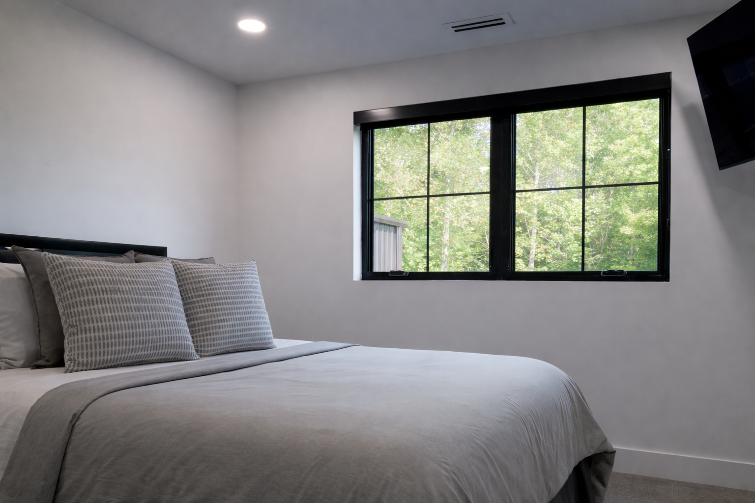 Minimal bedroom with a bed, gray bedding, and a black-framed window overlooking greenery