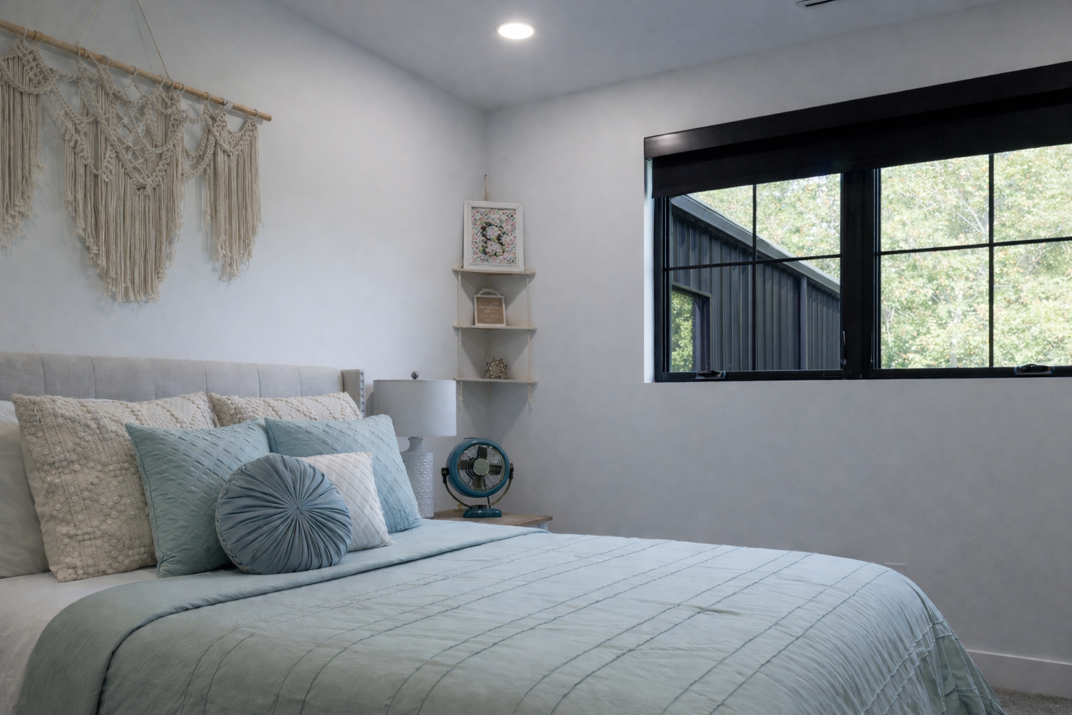 Bright bedroom with a bed, soft blue bedding, wall decor, and a large window overlooking greenery