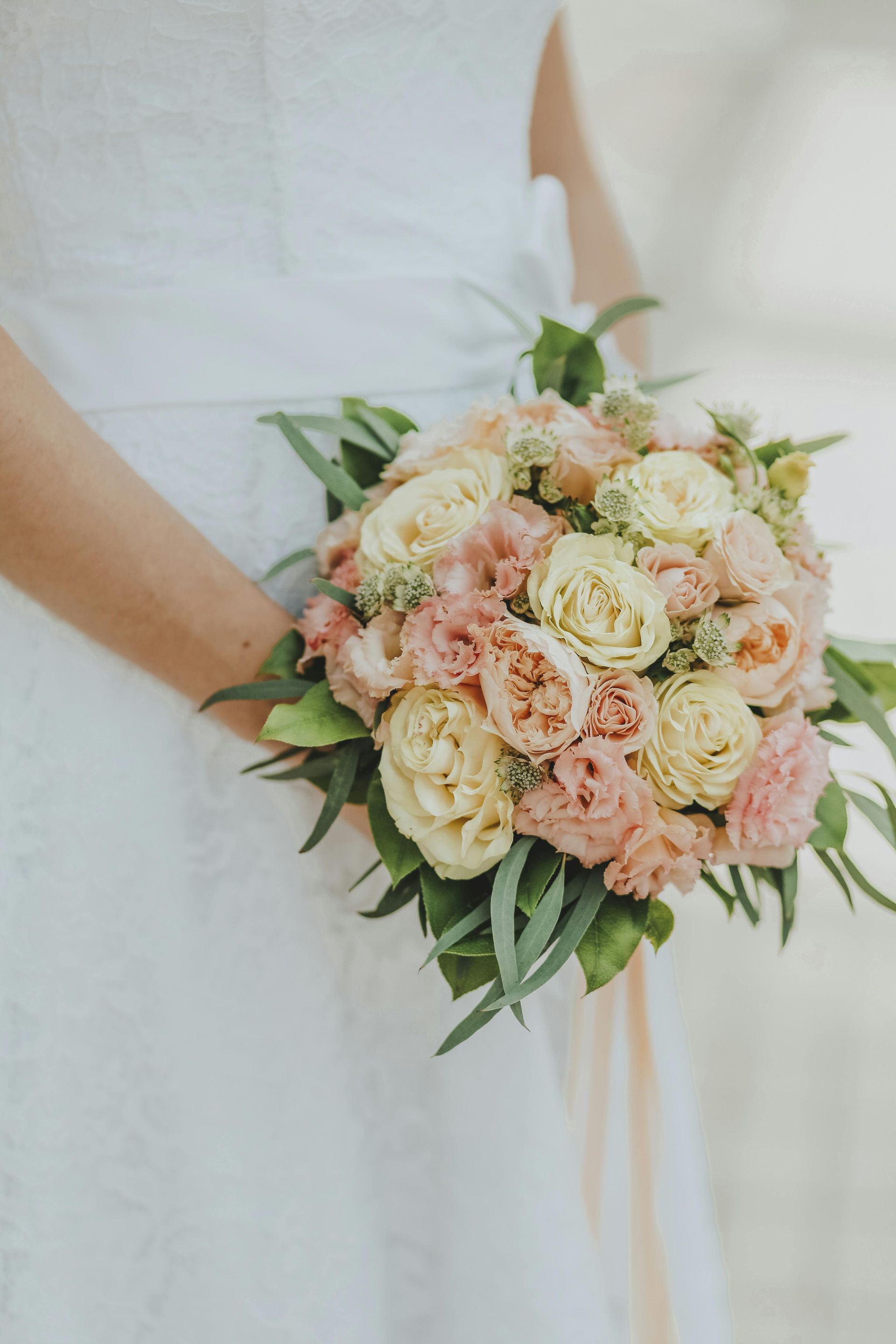 Person holding a bouquet of pale pink and cream flowers with greenery against a white dress
