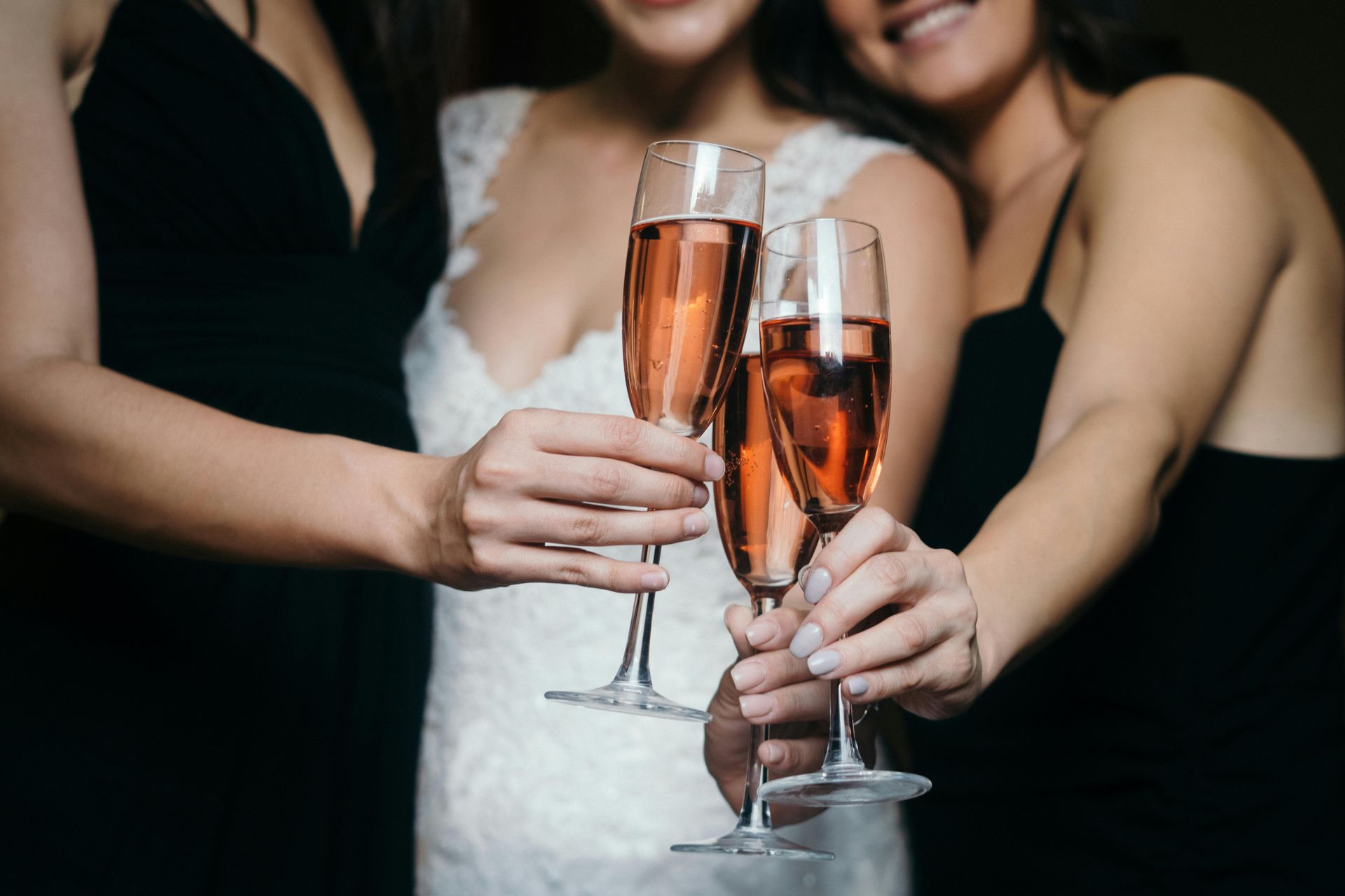 Hands clink champagne flutes at a party, with a woman in a white dress centered in the background.