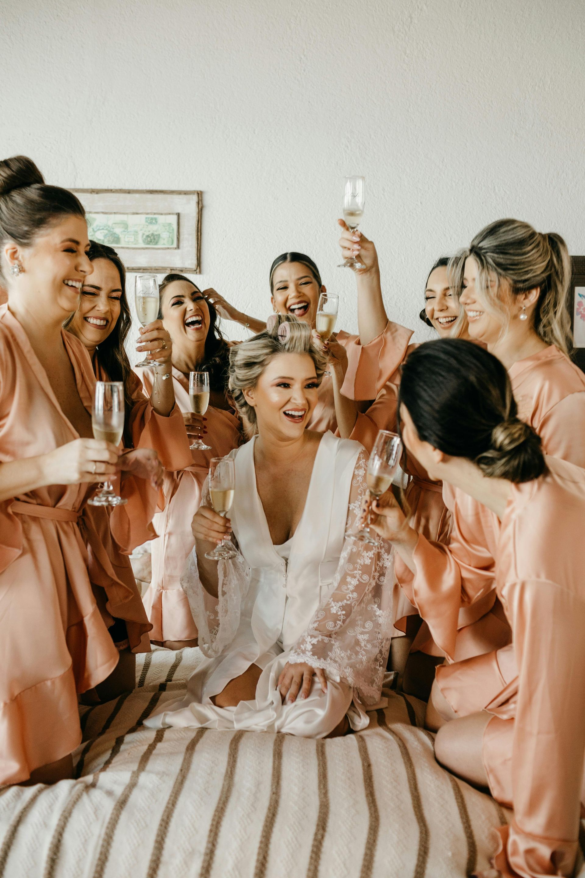 Bride surrounded by smiling bridesmaids in pink robes, raising champagne glasses in a celebratory room.
