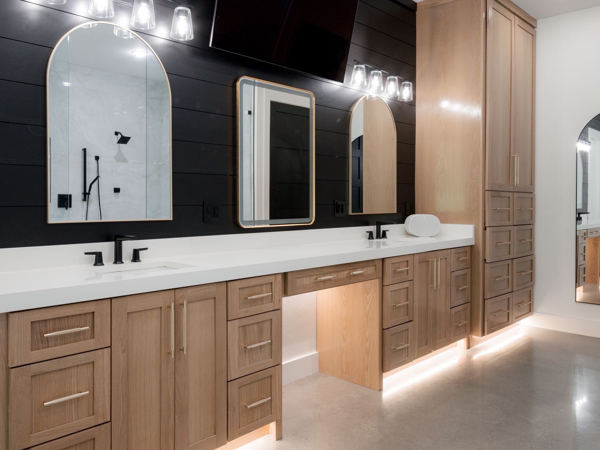 Modern restroom vanity with wood cabinets, white countertop, arched mirrors, and soft under-cabinet lighting