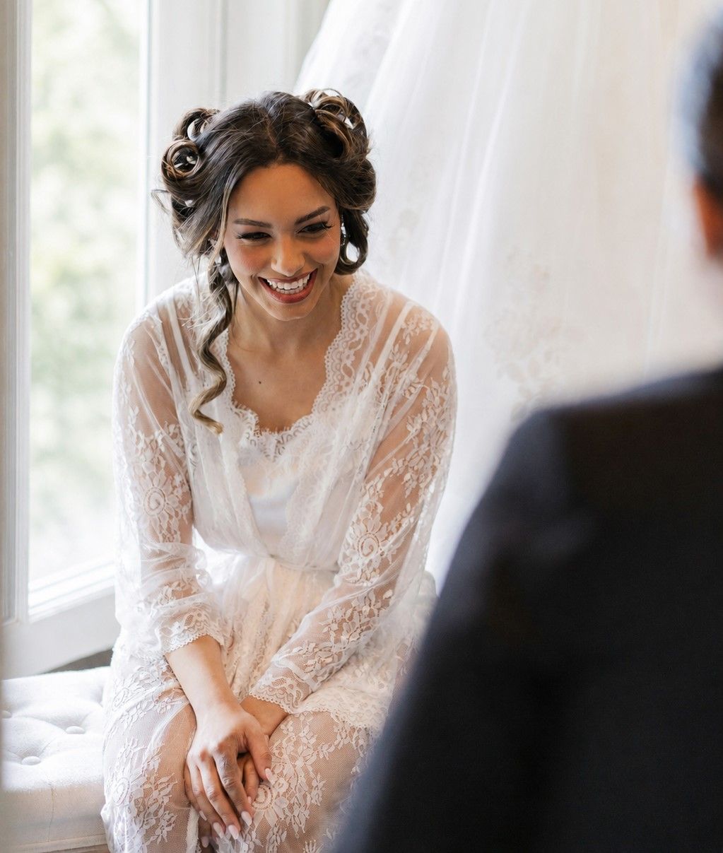 Smiling bride in a white lace dress seated near a bright window