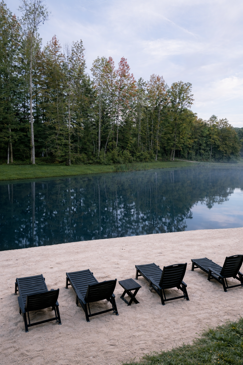 Wooden lounge chairs on a sandy shore beside a calm blue lake with a tree-lined forest beyond
