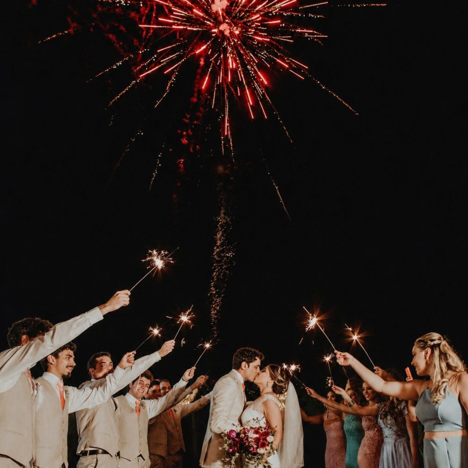 Wedding couple and guests raising sparklers under red fireworks at night