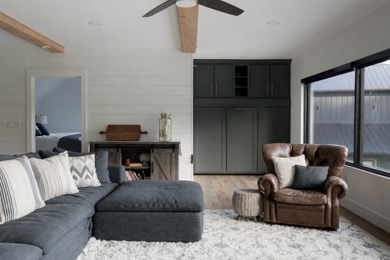 Bright living room with gray sectional, plaid armchair, fireplace, ceiling fan, and large windows