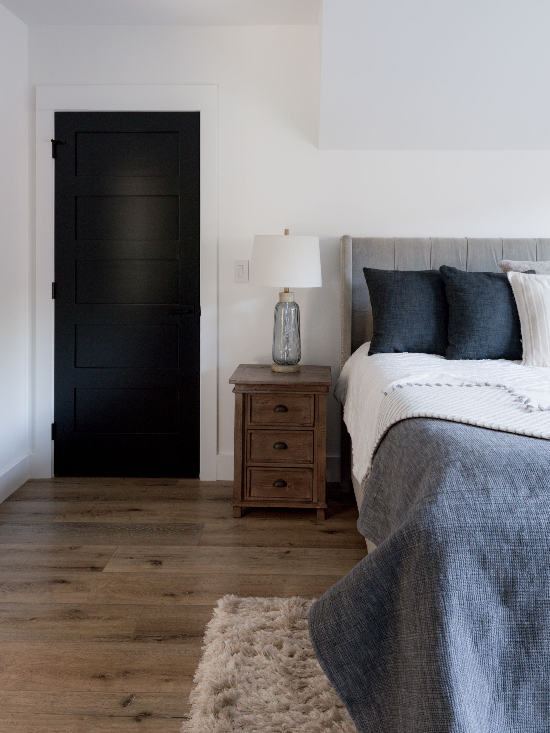 Minimal bedroom with black door, wooden nightstand, lamp, and gray bedding on a wooden floor.