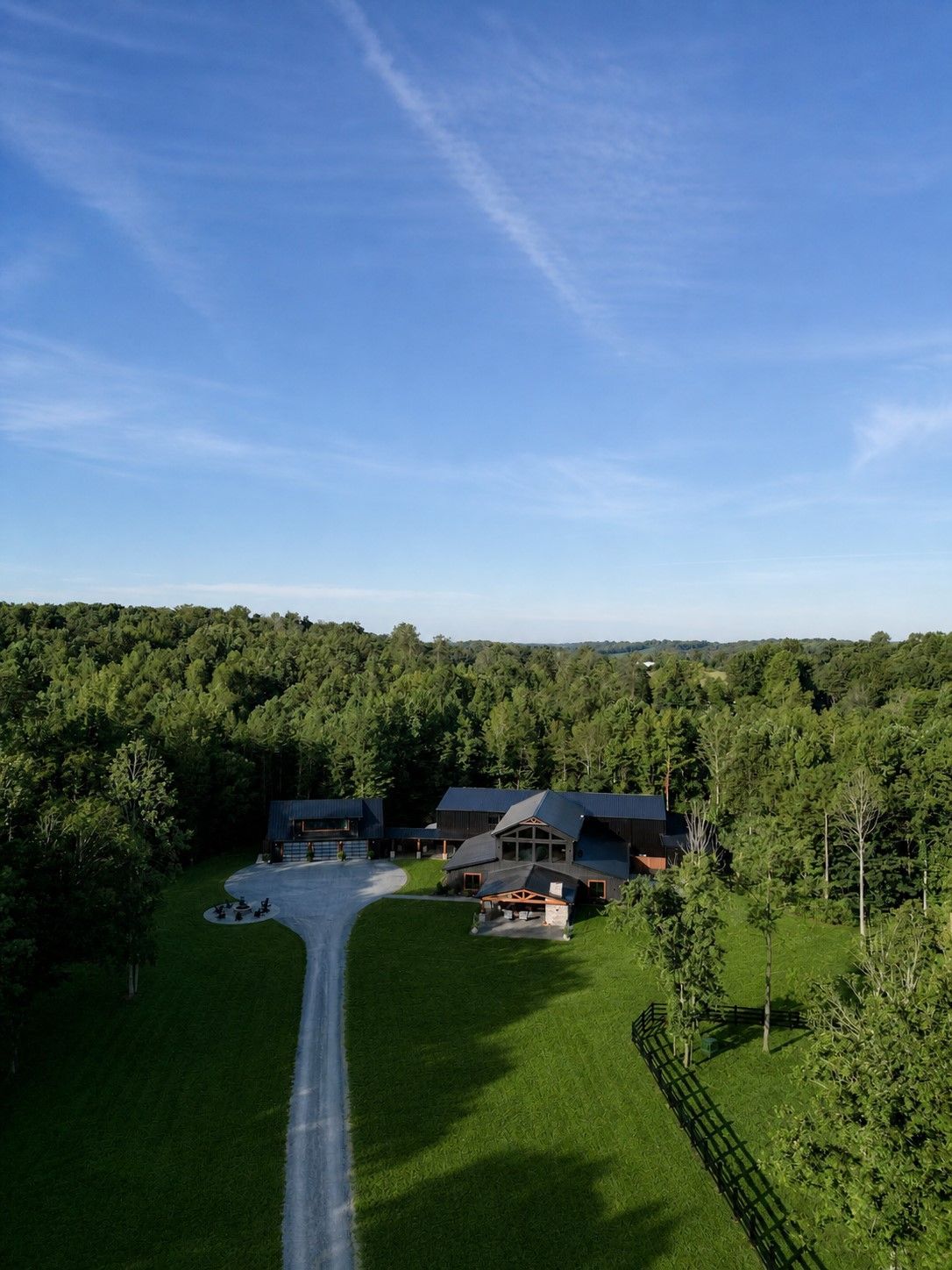 Aerial view of a long driveway leading to a house and outbuildings surrounded by green trees and open lawn