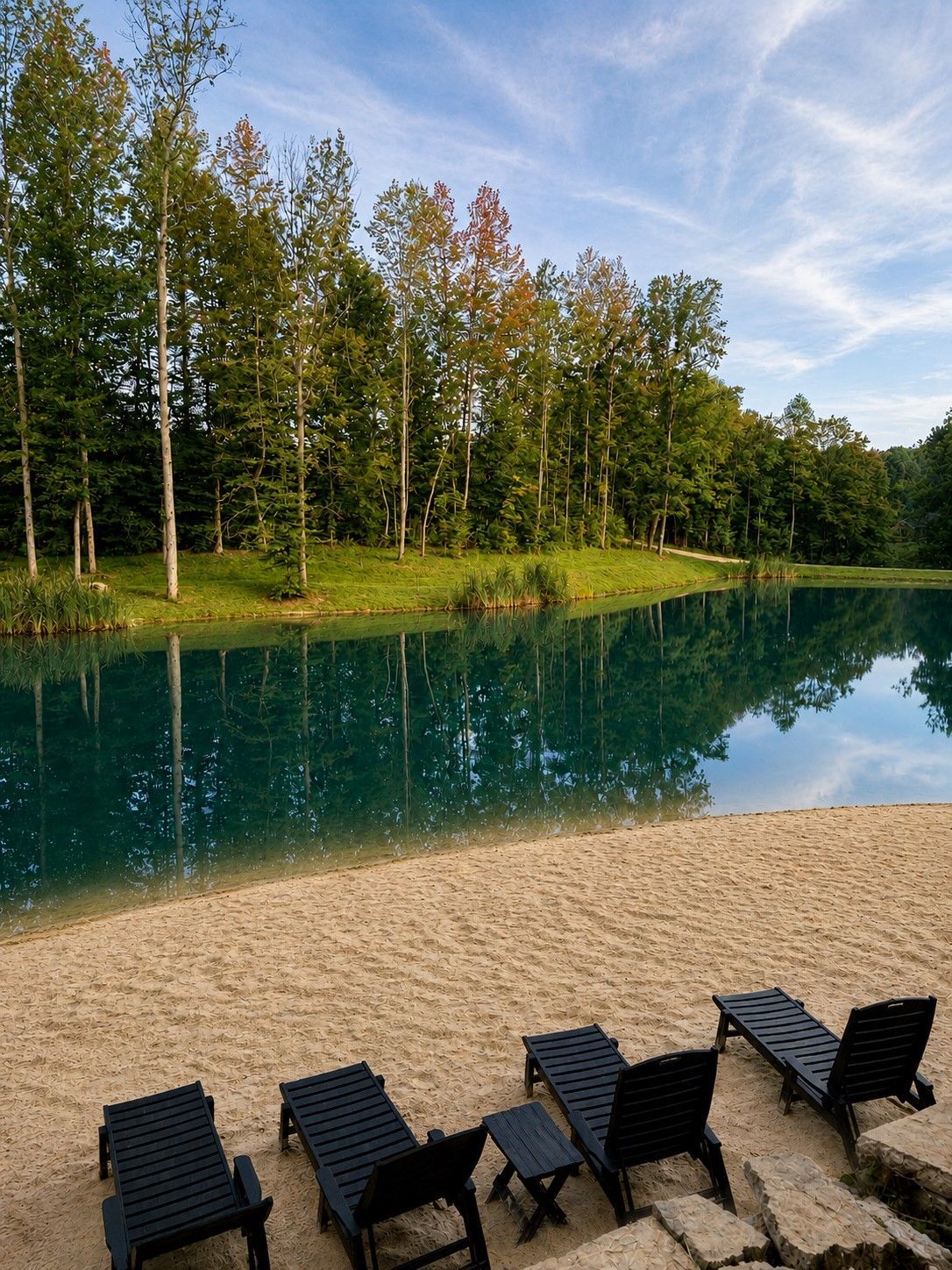 Chairs on a sandy shore beside a calm blue lake, with green trees reflected in the water.
