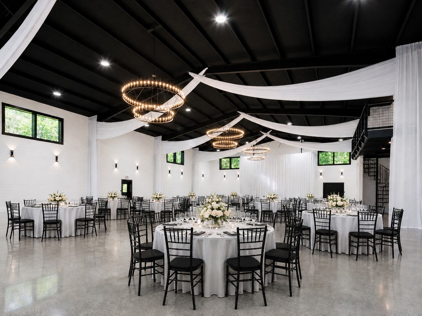 Elegant banquet hall with white tables, black chairs, black ceiling drapes, and ring chandeliers
