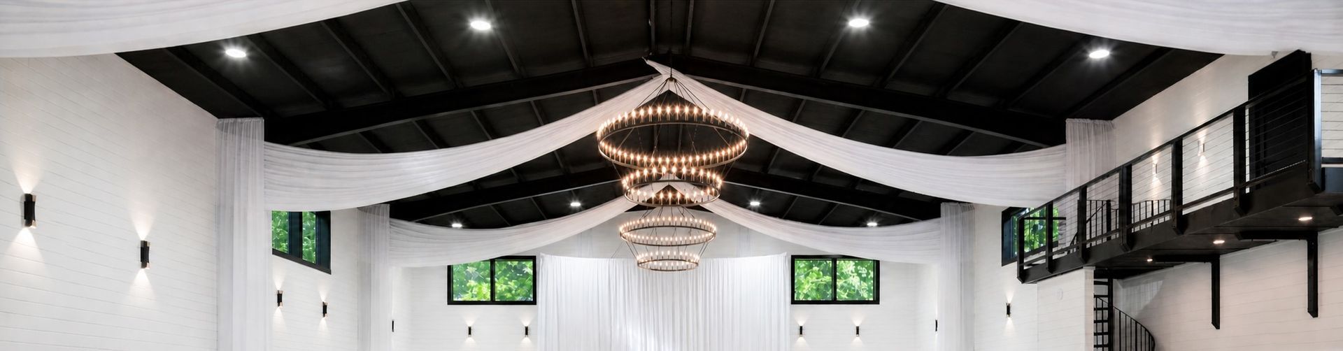 Wide white hall with black ceiling beams, hanging chandelier, and green exit signs overhead