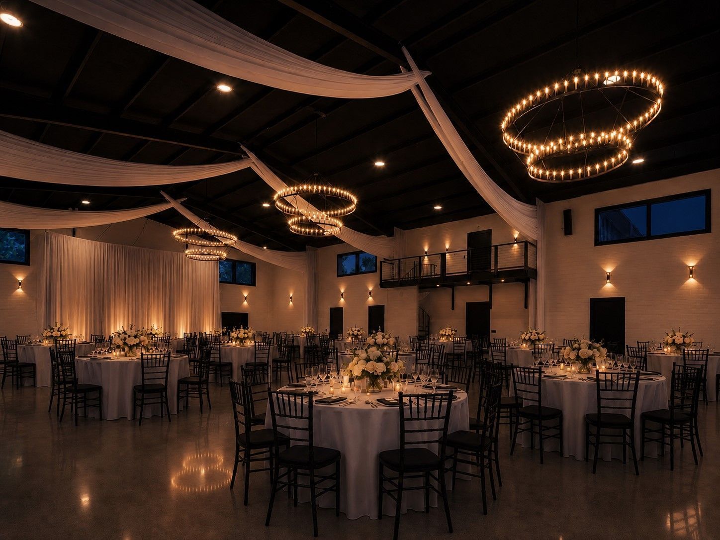 Elegant banquet hall with round tables, black chairs, warm chandeliers, and white draped walls