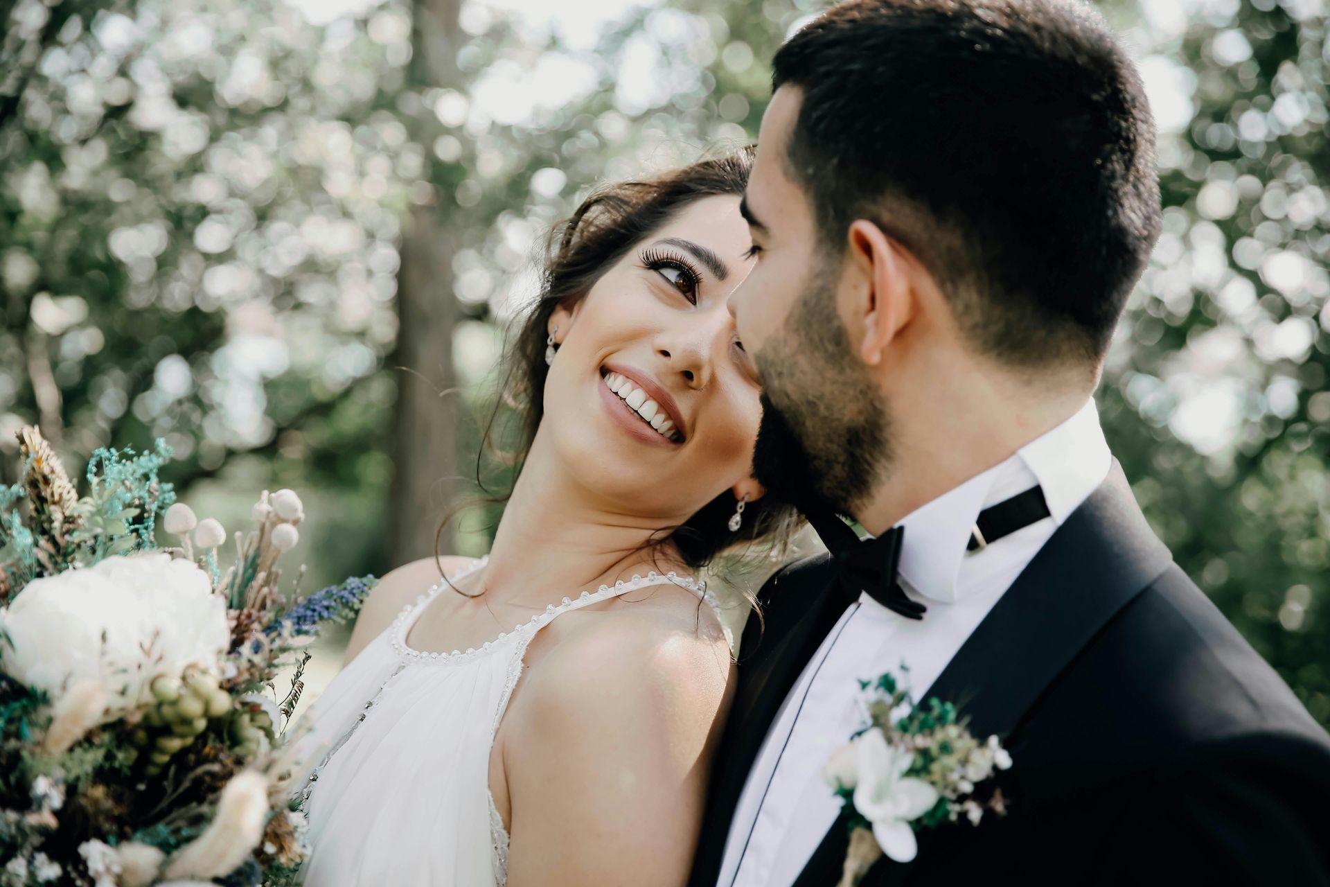 Smiling couple in wedding attire outdoors, gazing at each other with a bouquet in hand.