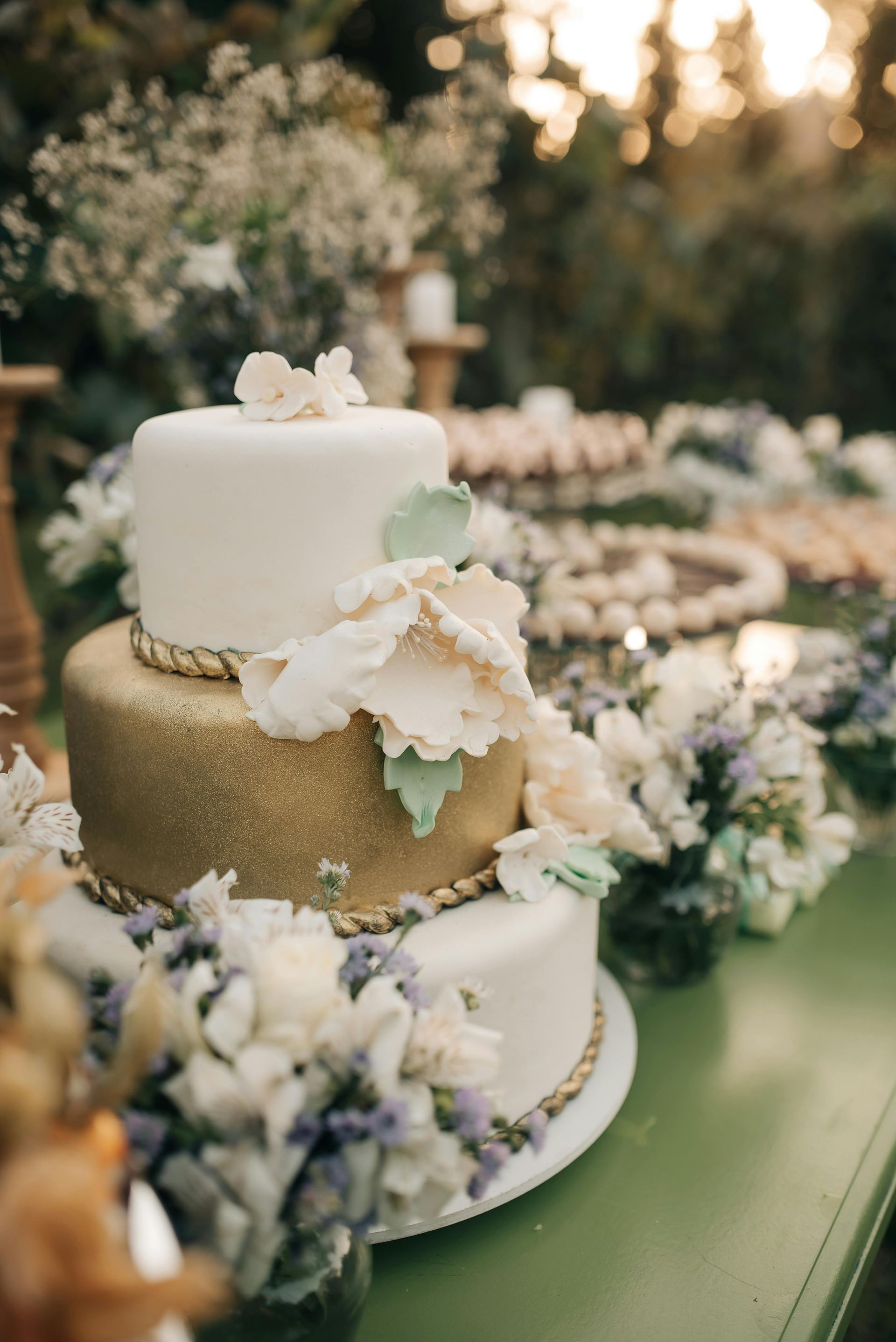 Elegant white-and-gold tiered wedding cake with floral decorations on a green table outdoors