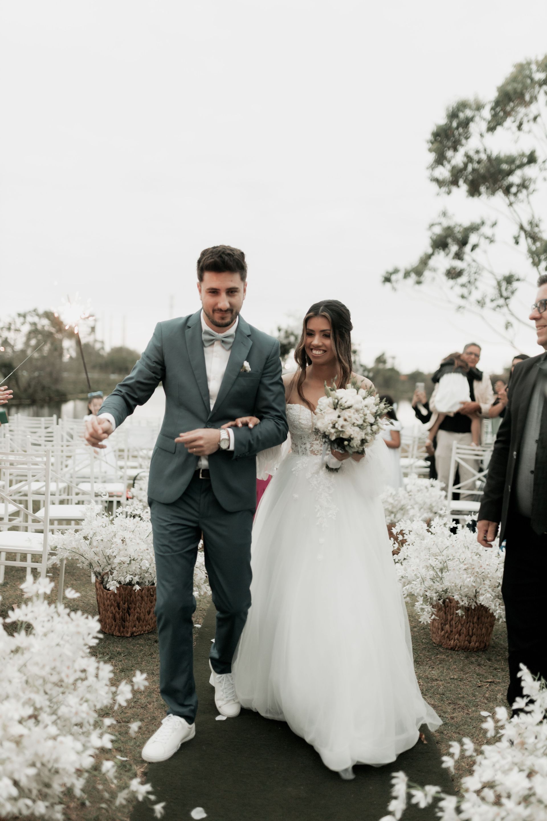 Newlyweds walking down a white-flower-lined aisle, holding hands and smiling outdoors
