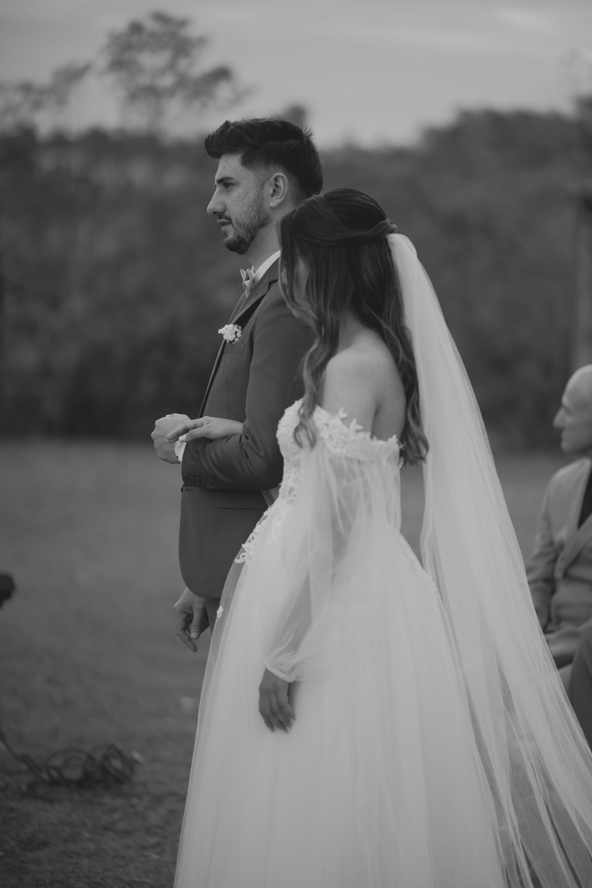 Bride in a white wedding gown and veil stands beside a groom in a suit outdoors.