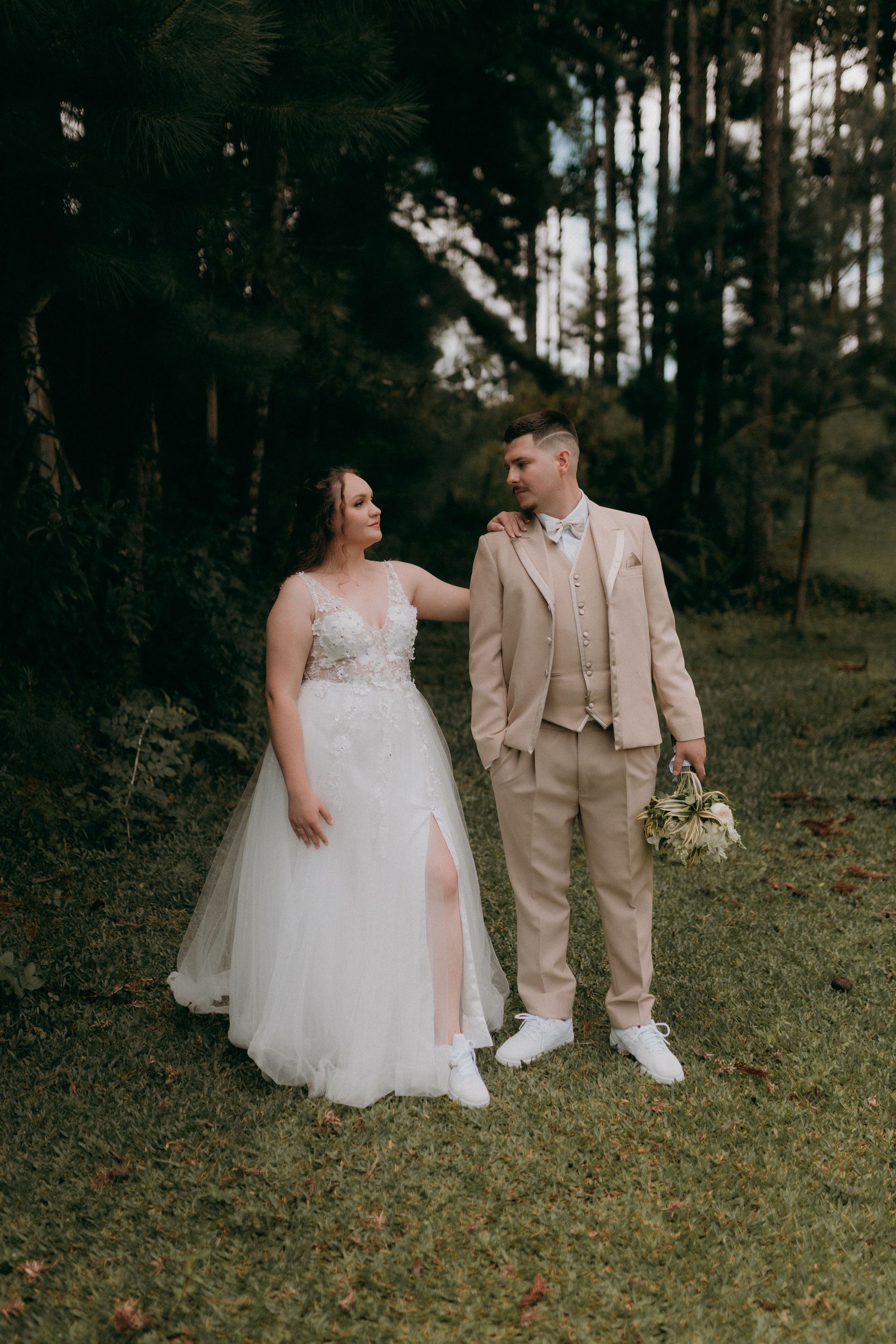 Bride and groom standing together outdoors in a wooded garden, holding hands and flowers