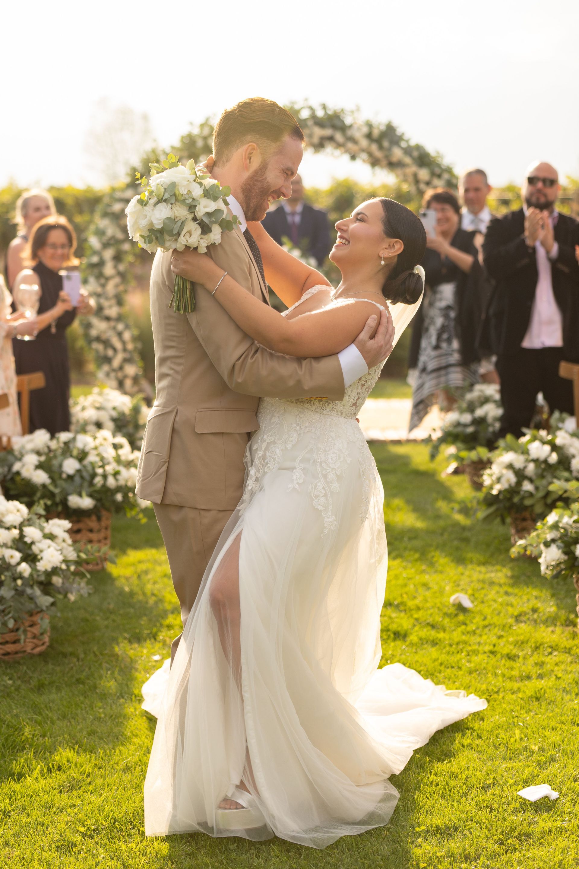 Wedding couple dancing outdoors in sunlight, bride in white gown and groom in tan suit, surrounded by guests