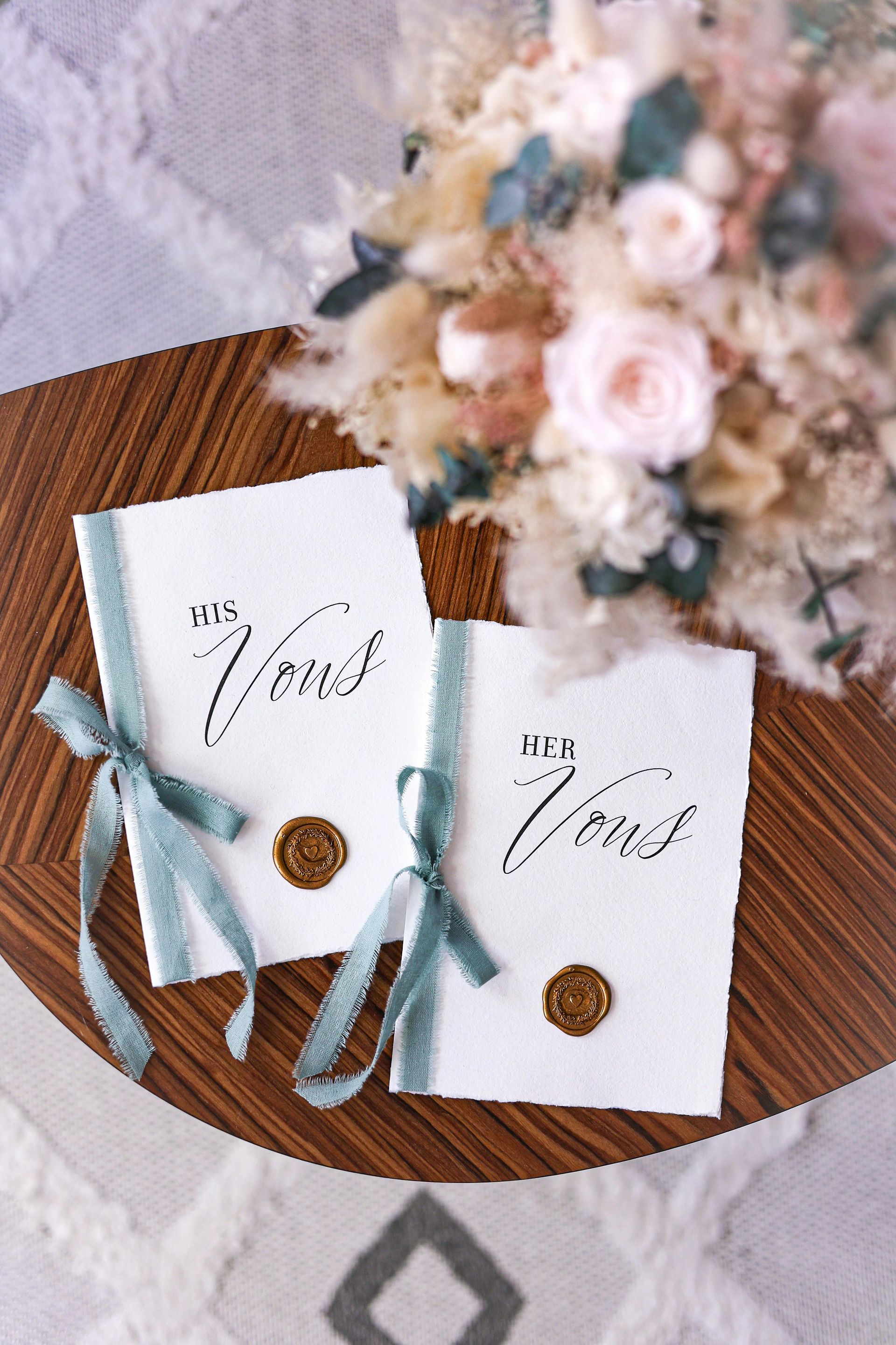 Wedding invitation cards on a wooden table with teal ribbon and gold seal, beside a pastel floral bouquet