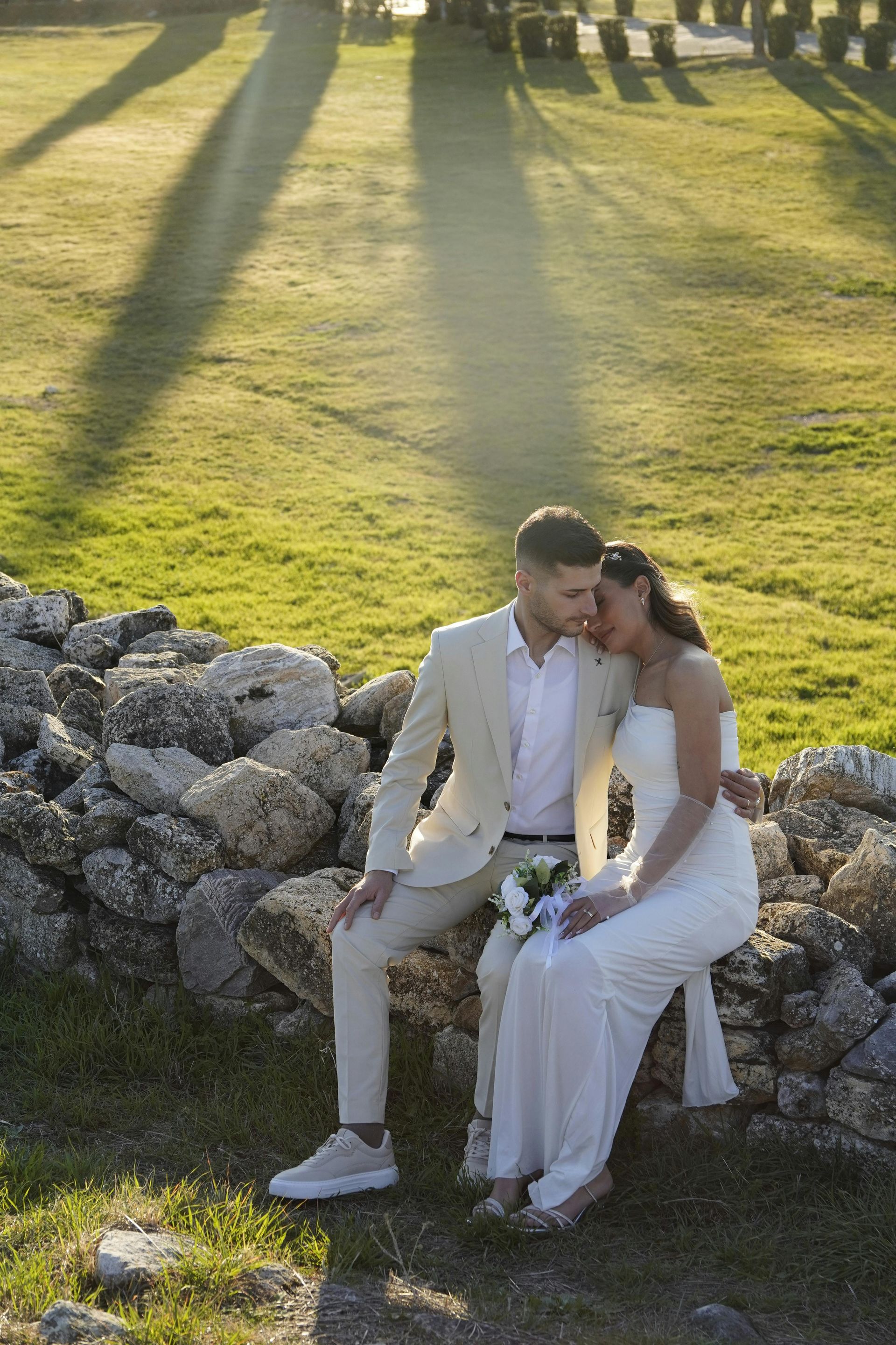 Couple in white wedding attire sitting on a stone wall in a sunlit garden