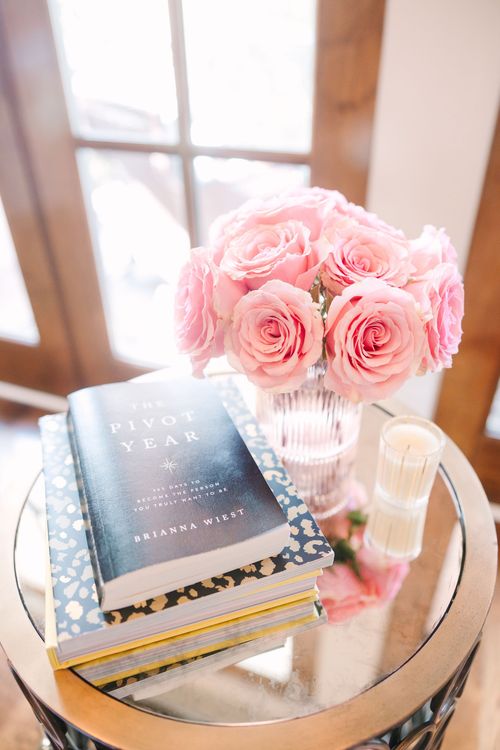 A vase of pink roses is sitting on a glass table next to a stack of books.