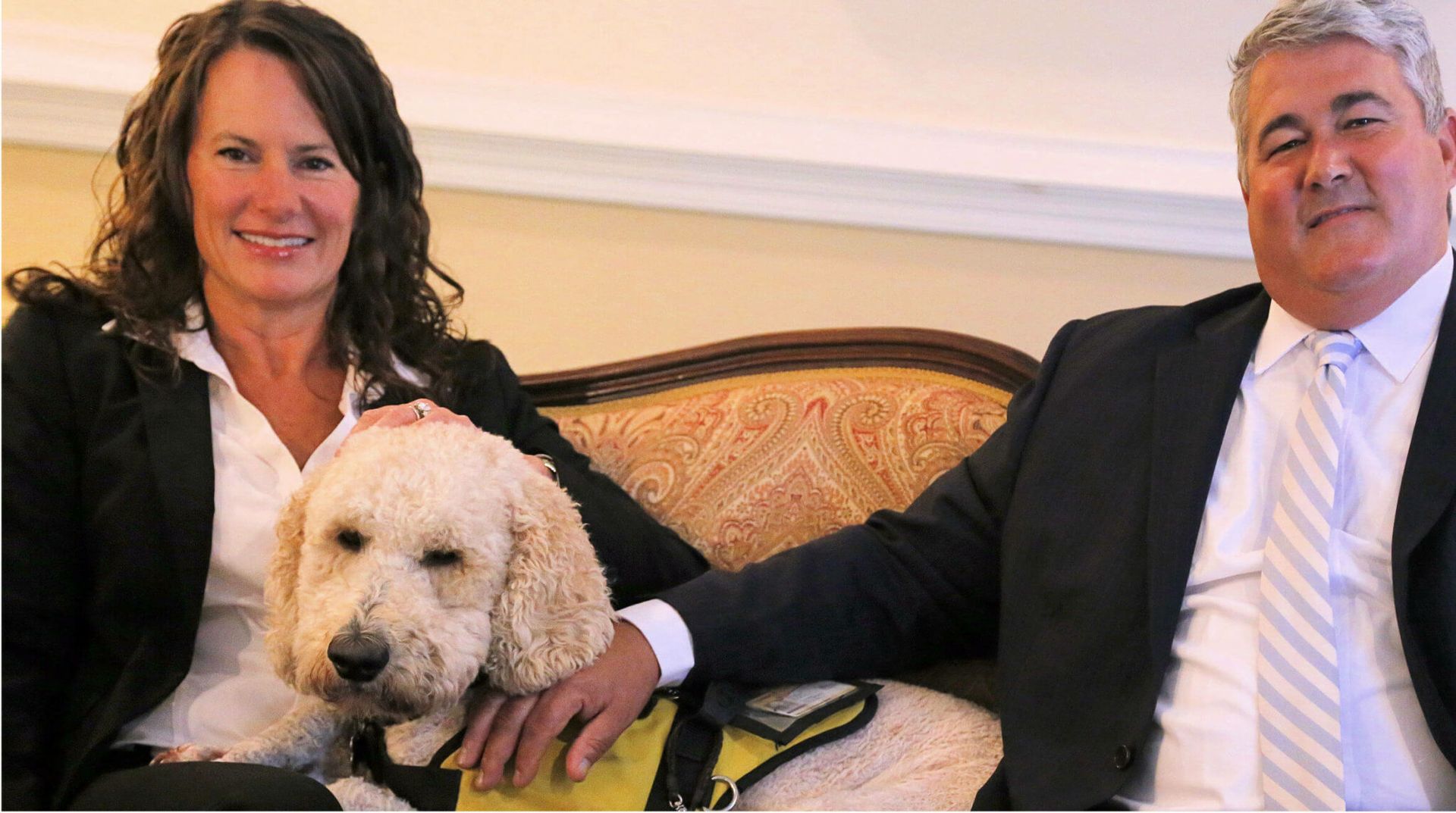 Two people seated on a couch with a curly white dog between them in a formal living room.