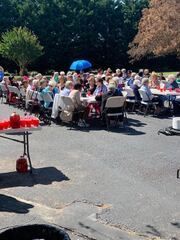 People seated at long tables outdoors for a gathering, with a blue umbrella and trees in the background.