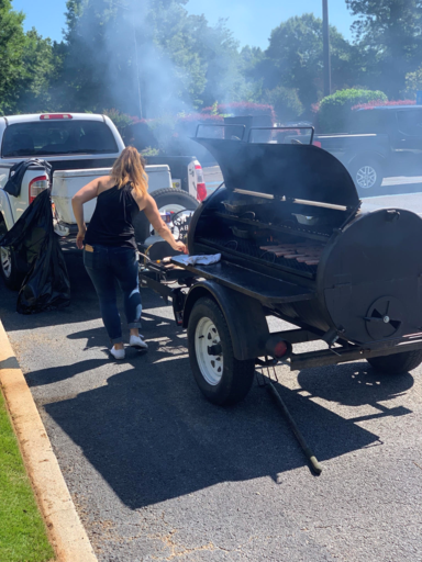 Woman tending a black smoker grill in a parking lot, with smoke rising behind parked vehicles.