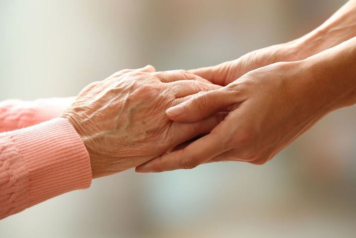 Two hands gently holding in a caring gesture, one elderly in a pink sweater and one younger hand