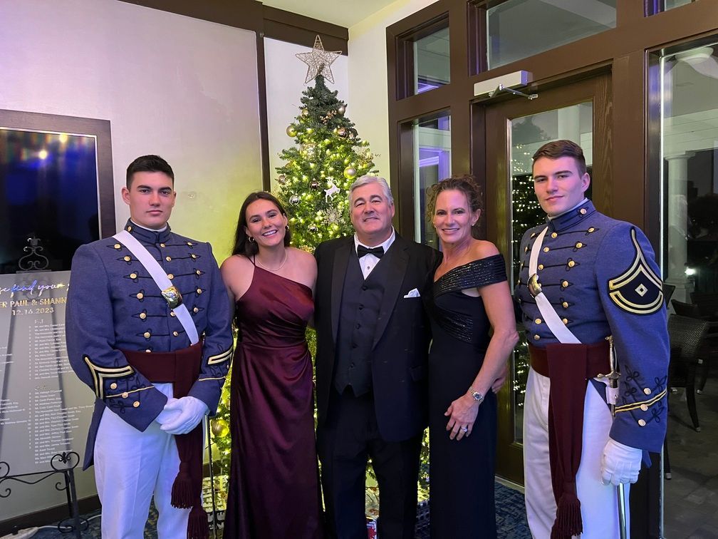 Five people pose indoors beside a decorated Christmas tree, with two in formal blue uniforms.