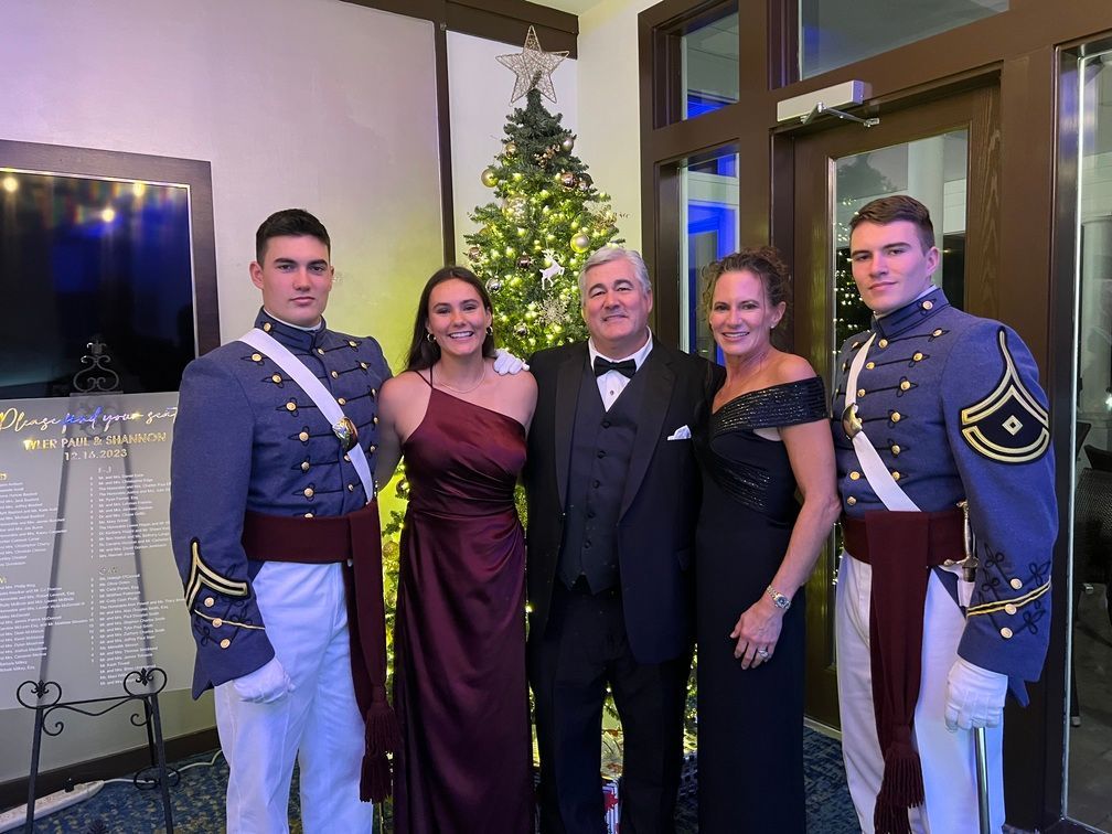Five people posing indoors by a decorated Christmas tree, two in blue dress uniforms and three in formal attire