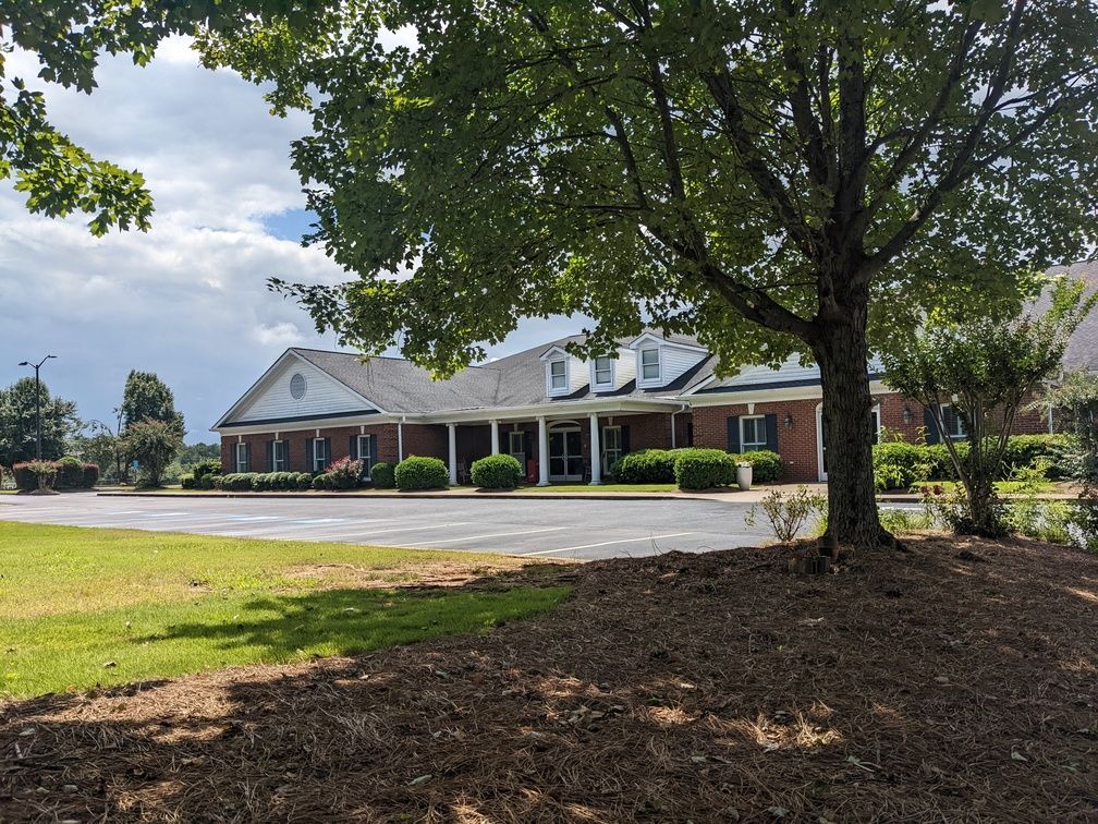 Brick building with white trim and covered porch behind a grassy lawn and large shade trees