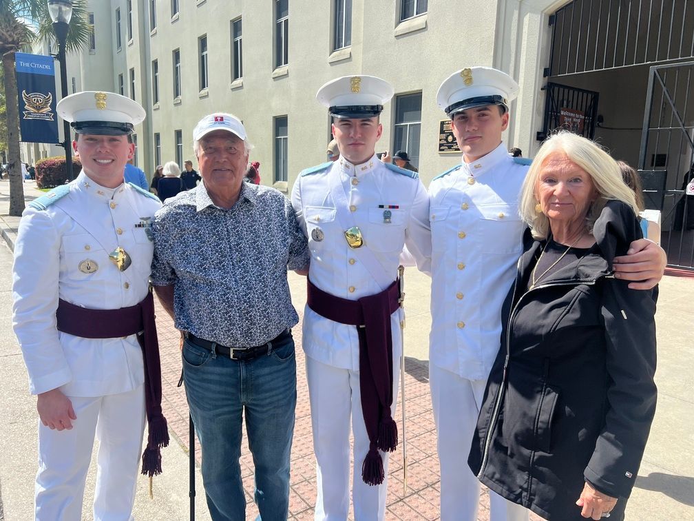 Four people pose outdoors beside a building, with two in white naval uniforms and two in casual clothes.
