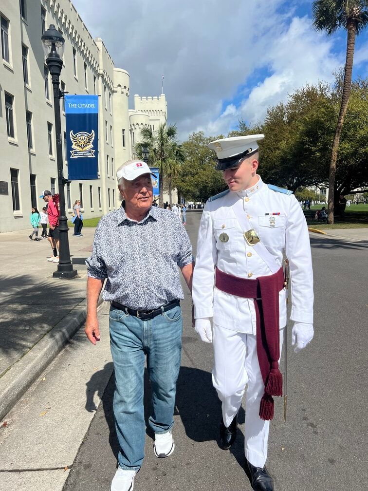 Man in a white naval uniform walks beside a man in a cap on a sunny street near a stone building.