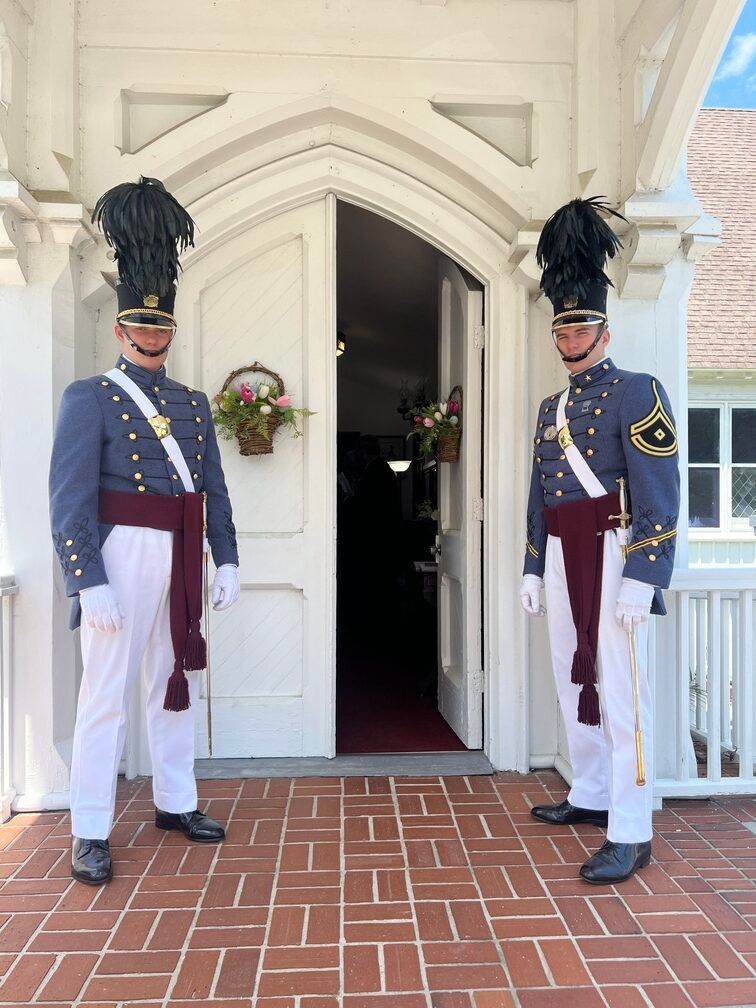 Two uniformed guards standing at a white church doorway, wearing tall black hats and maroon sashes.