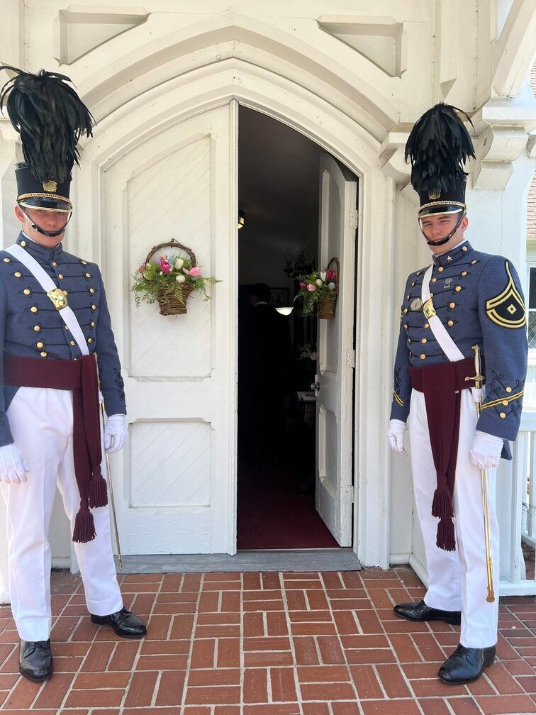Two ceremonial guards standing at a white doorway with wreaths, red brick floor beneath.