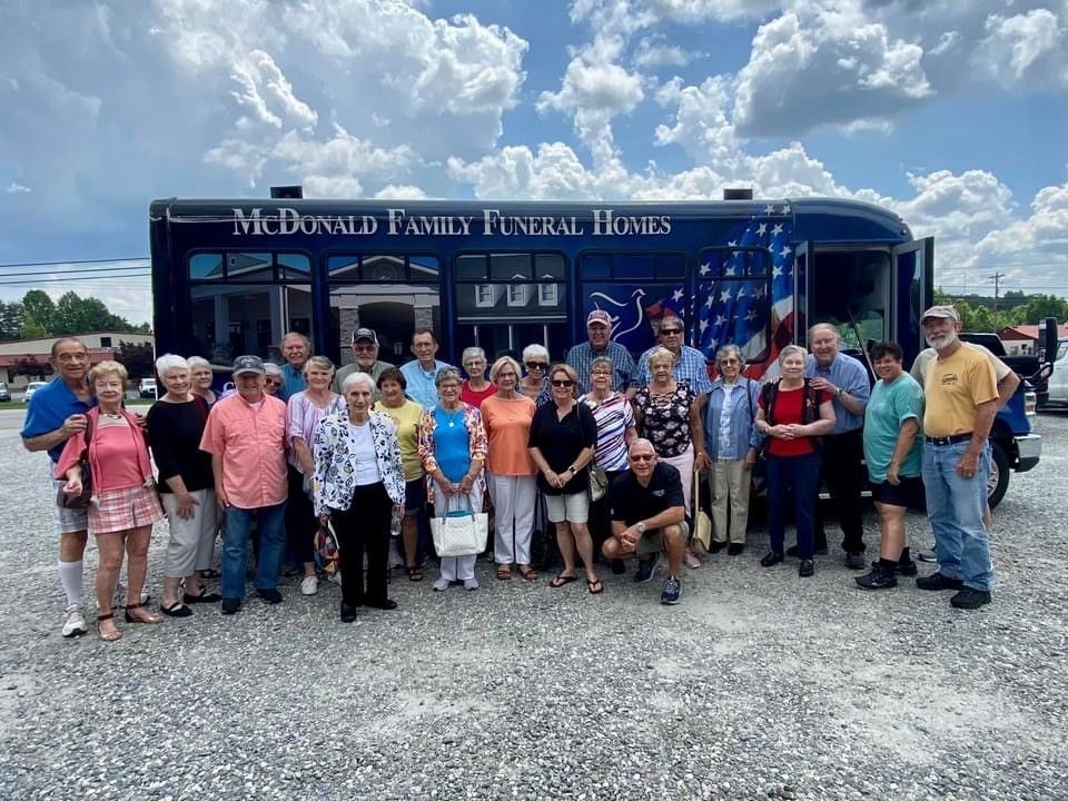 Large group posing in front of a blue funeral home shuttle under a cloudy sky.
