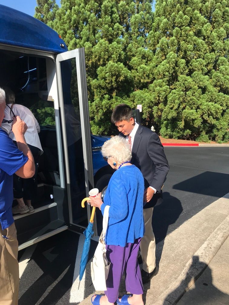 Woman with cane boarding a bus with assistance from a man, beside another passenger at the open door.