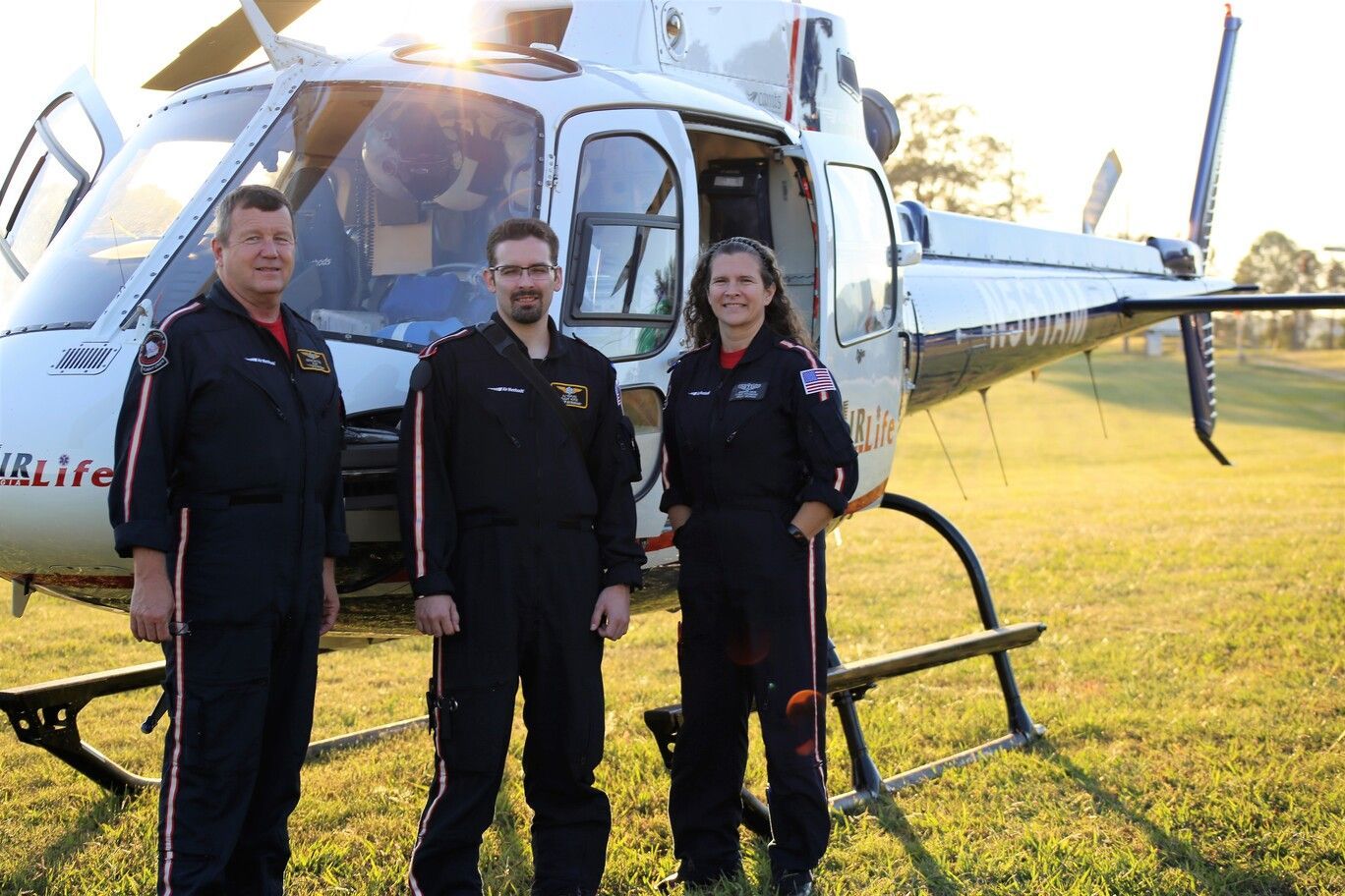 Three people standing in front of a white helicopter on a grassy field at sunset.