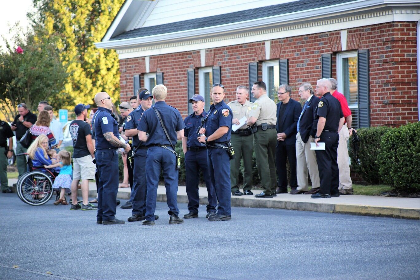 Police officers and men gather outside a brick building near a wheelchair user and parked car.