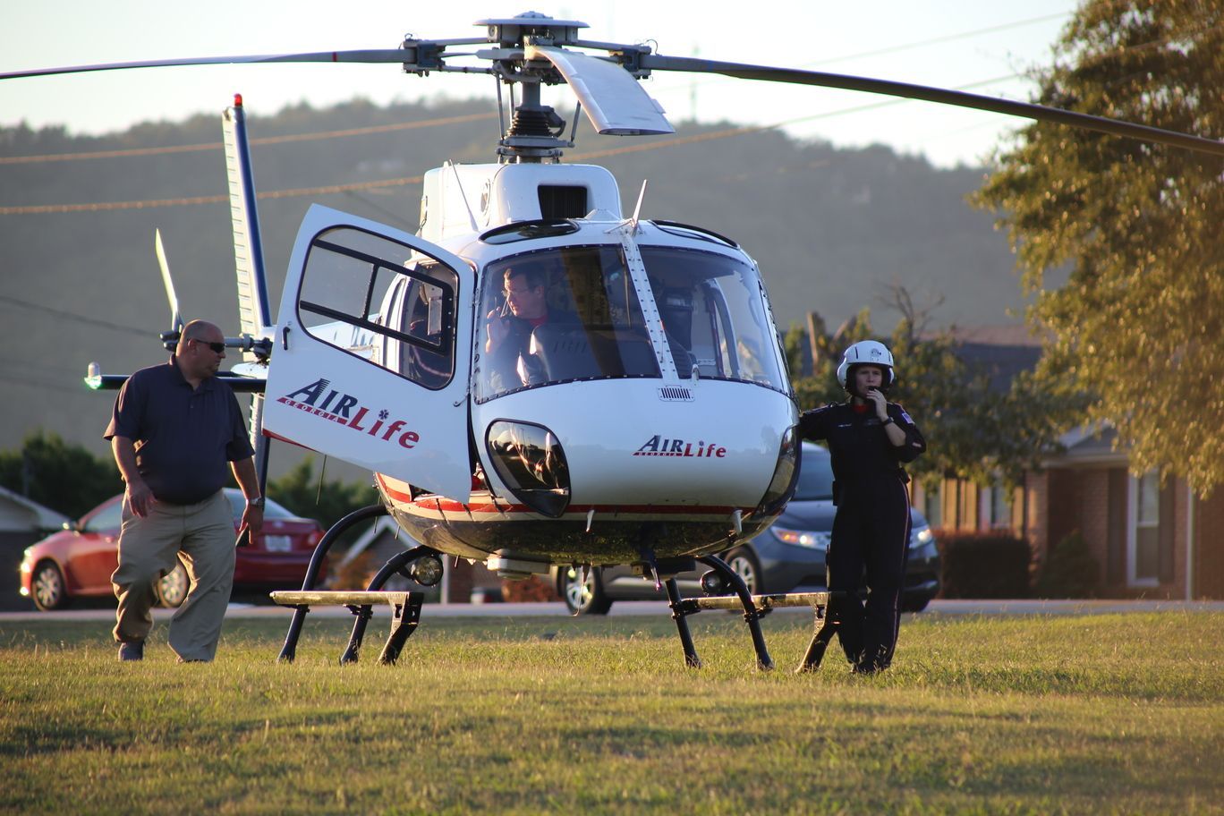 Helicopter on a grassy field with two people nearby, rotors spinning, at sunset