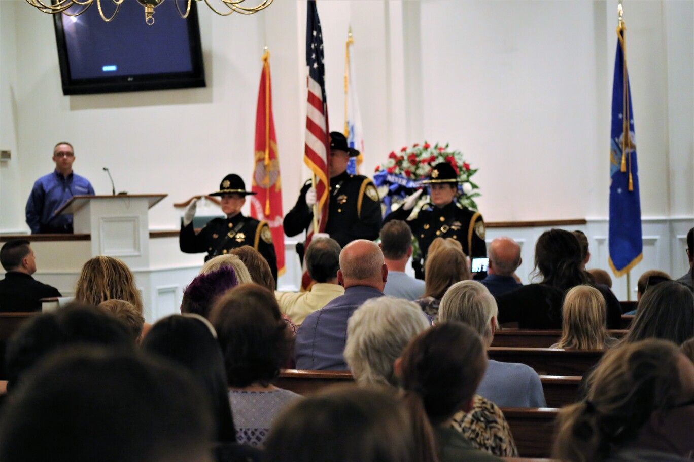 Audience watching a ceremony with speakers at a podium and flag display in a hall.