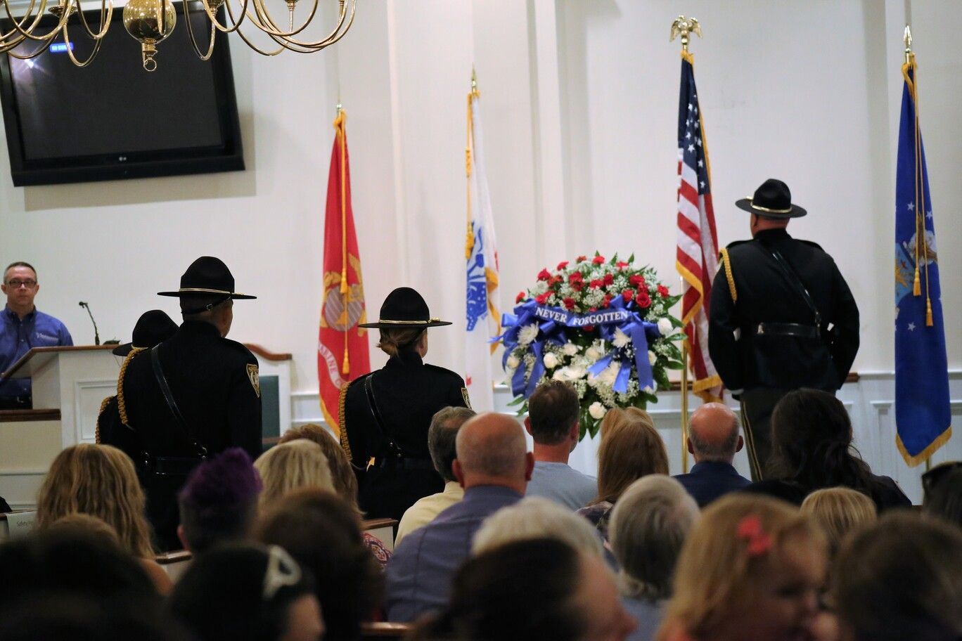 Indoor memorial service with uniformed officers standing before floral wreaths and flags, audience seated in front.