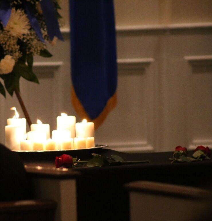 Lit candles on a table in a dim ceremonial room with flowers and a blue banner in the background