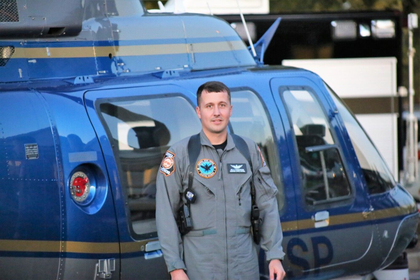 Pilot in gray flight suit standing beside a blue helicopter in a hangar