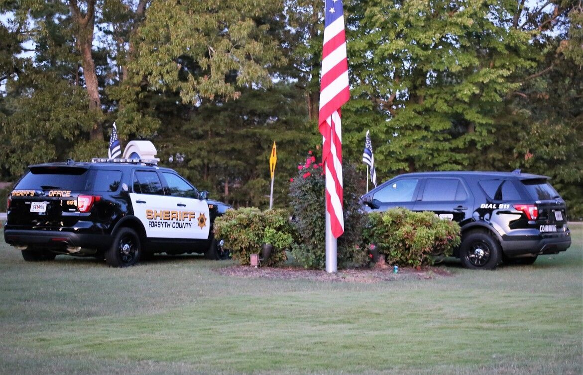 Two police SUVs parked on a grassy lawn beside a large American flag and trees.