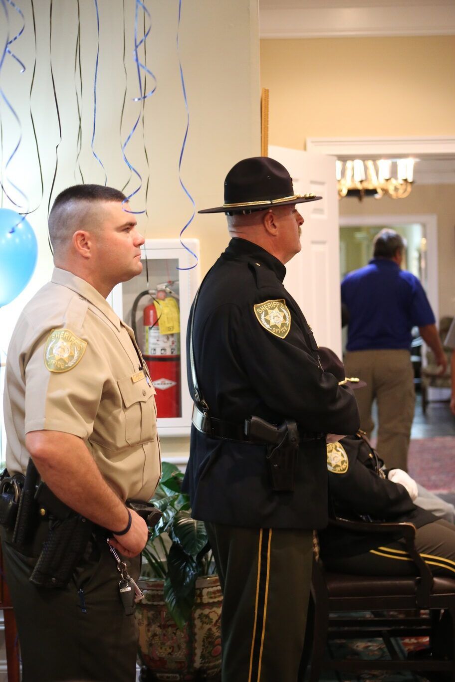 Two uniformed officers stand indoors near a hallway decorated with blue streamers.